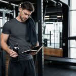 Lifter reviewing a workout notebook beside a barbell rack and treadmill for exercise and weight loss planning