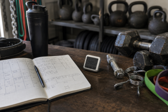 Training program planning setup with dumbbells, bands, lifting belt, timer, water bottle, and workout notebook