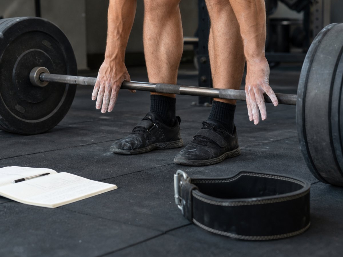 Deadlift setup close view with bar over midfoot, chalked hands, belt, and training log