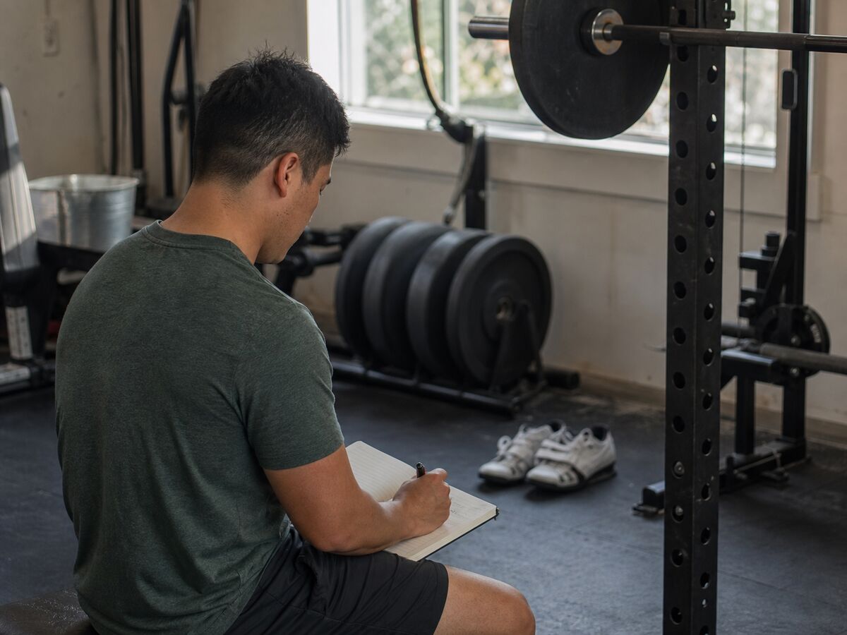 Lifter recording a gym session in a notebook beside a squat rack