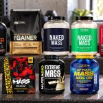 Ten mass-gainer packages arranged on a gym bench, including Naked dairy and vegan tubs and two different BSN formulas