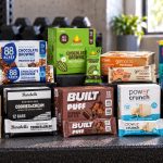 Ten protein bar products arranged on a gym bench with branded boxes and individual wrappers facing forward