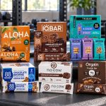 Ten protein bar brands arranged on a gym bench with boxes and individual wrappers facing forward