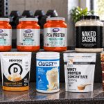 Ten baking-suitable protein powder packages with visible labels arranged on a gym bench