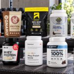 Ten sport-certified protein powder packages arranged on a gym bench with readable front labels