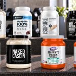 Eight protein powder packages with visible front labels arranged on a gym bench